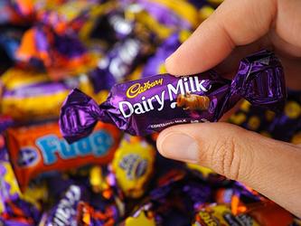 Hand holding small bar of Cadbury dairy milk, with other Cadbury wrapped chocolates in the background